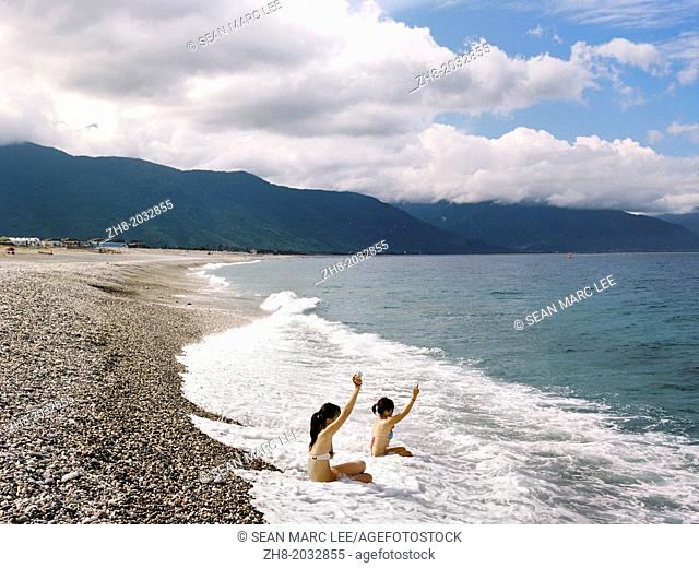 Two young women in bikinis sit on a rocky beach while holding their cameras to the sky at Chi Shing Tan in Hualien, Taiwan