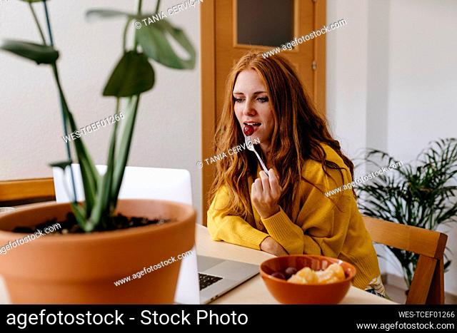 Young woman eating fruit while using laptop at home