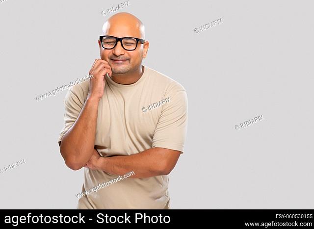 Portrait of a bald man wearing eyeglasses smiling while thinking something with closed eyes against plain background