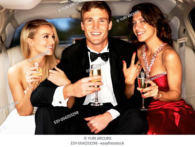 A handsome young celebrity drinking champagne flanked by two gorgeous woman in a limousine
