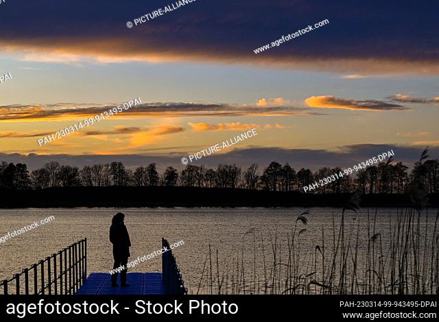 13 March 2023, Brandenburg, Altfriedland: In the backlight of the setting sun, a woman stands on a jetty at Klostersee lake in the Märkisch-Oderland district