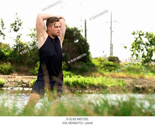 Young man exercising beside river, stretching arms