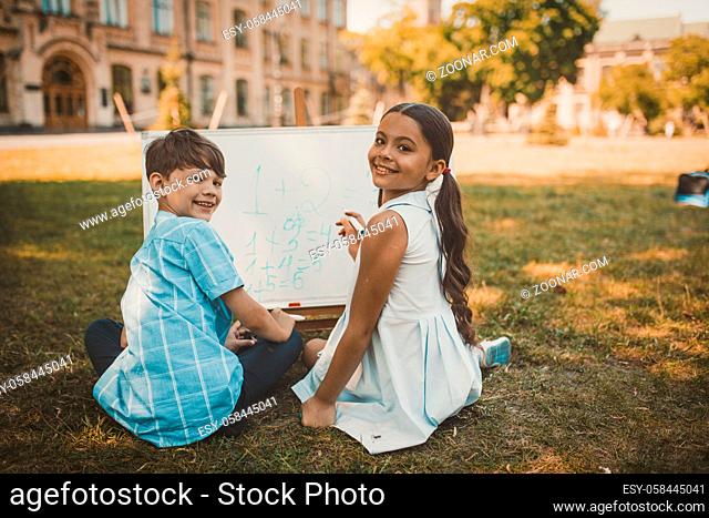 Happy Boy And Girl Smiles Looking At Camera, Cheerful Children Look Back Sitting On Grass And Writing Numbers By Color Markers On Whiteboard, Toned Image