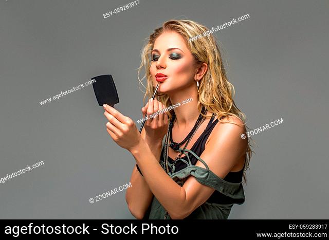 beautiful blond young woman with long blond hair holding pocket mirror and applying makeup. studio beauty shot on grey background. copy space