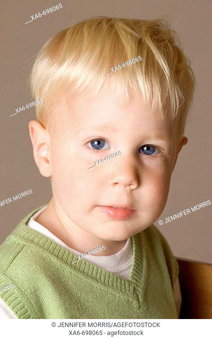 A blonde-hair, blue-eyed two year old boy looks straight at the camera. He is wearing a white t-shirt and green jumper