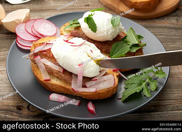 sandwich on toasted white slice of bread with poached eggs, green leaves of arugula and radish, morning breakfast on a gray wooden board, close up