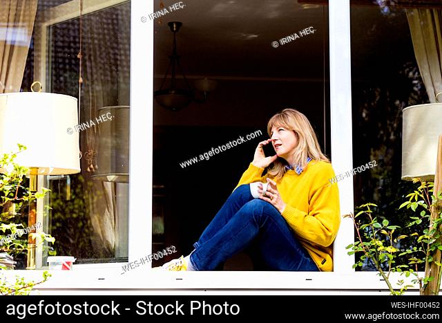 Mature woman talking on smart phone while sitting in window sill at home