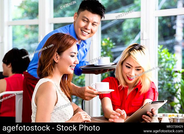 Smiling Asian waiter serving two stylish ladies beverages in a restaurant as they browse the internet on a tablet pc