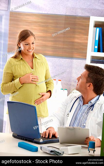 Pregnant woman standing in consulting room, looking at doctor at computer, smiling