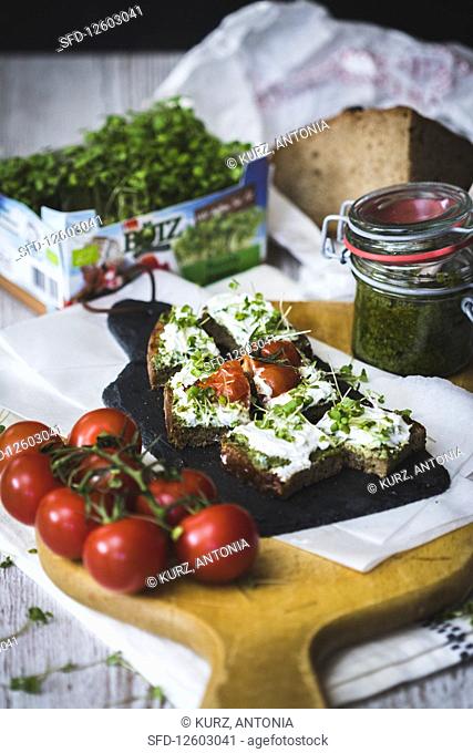 A slice of bread topped with cream cheese, cress and tomatoes with pesto in the background