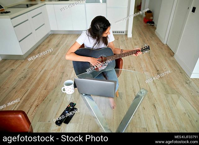 Mid adult woman practicing guitar while sitting with laptop at home