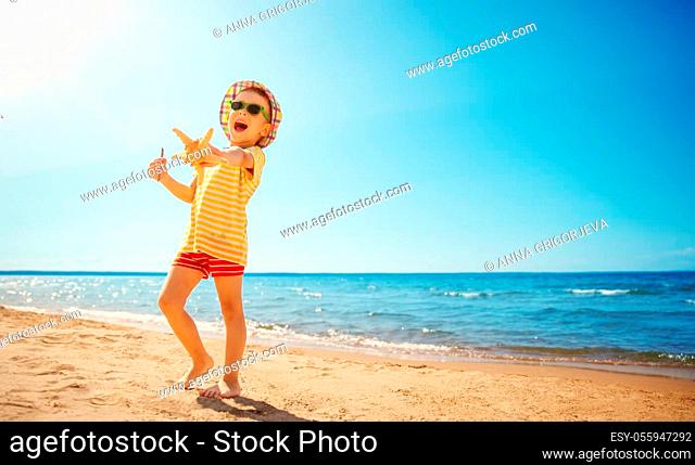 four years old boy smiling at the beach in hat with sunglasses. Child on vacations at sea. Seaside with clean sand, blue sky and sun in summer