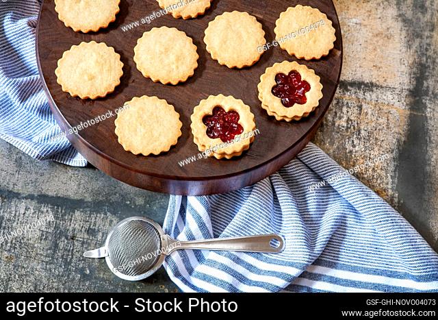 High angle view of Linzer tarts being assembled on round wood board with blue towel and mesh sieve