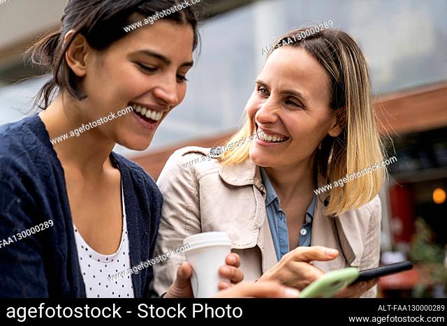 Low angle side view of two female freelancers working outdoors and having a good time while checking their smartphones