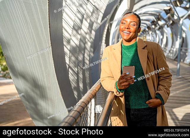Happy woman holding mobile phone standing with hand in pocket on bridge