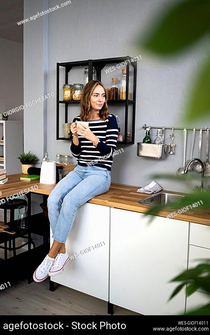 Smiling woman with mug sitting on kitchen counter