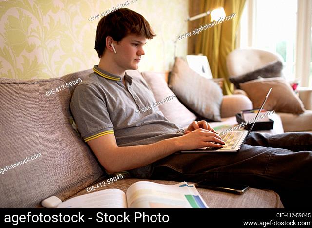 Teenage boy with earbuds using laptop on living room sofa