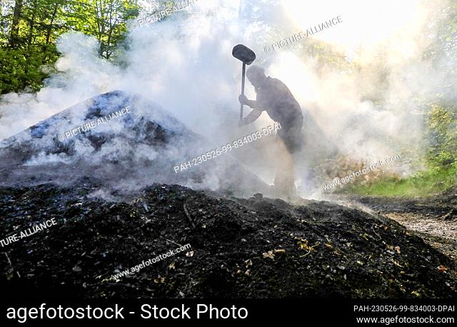 26 May 2023, Baden-Württemberg, Hayingen: Charcoal burner Max Geiselhart stands on a smoldering charcoal pile and compresses it with a wooden hammer
