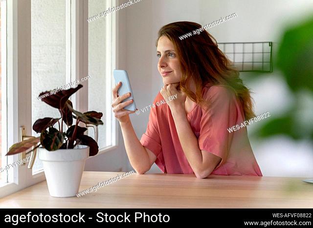 Thoughtful woman with hand on chin holding smart phone at home