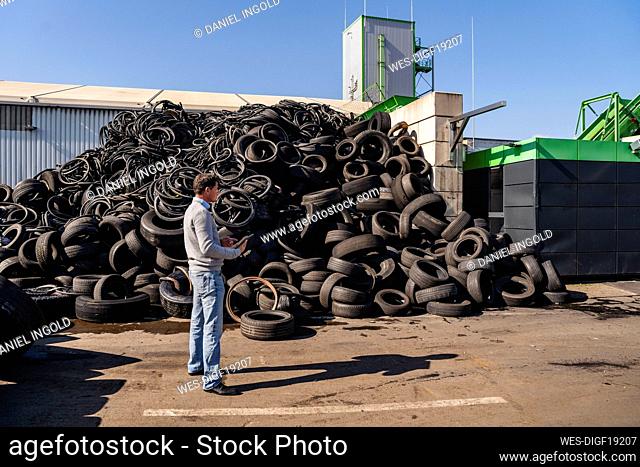 Mature businessman with tablet PC standing by heap of rubber tires at recycling plant