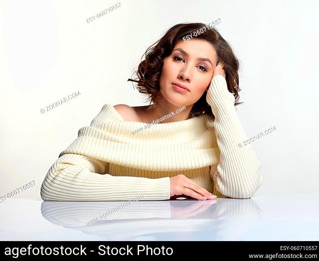 Beautiful brunette woman dressed in large white woolen sweater is sitting at white table on gray background. Hand running in hair and props head