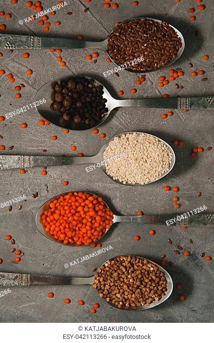 Spoons with different superfoods - flax seeds, sesame, pepper, red lentils, buckwheat on a gray background. Vertical photo