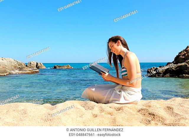 Relaxed tourist sitting on the sand of the beach reading a paper book on summer vacation