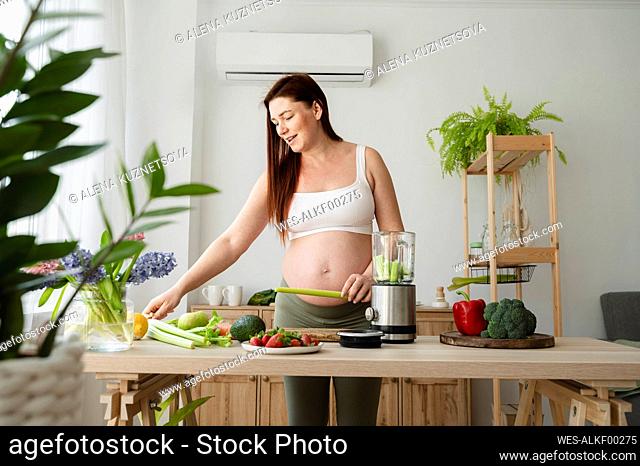 Pregnant woman making smoothie with vegetables on table in kitchen