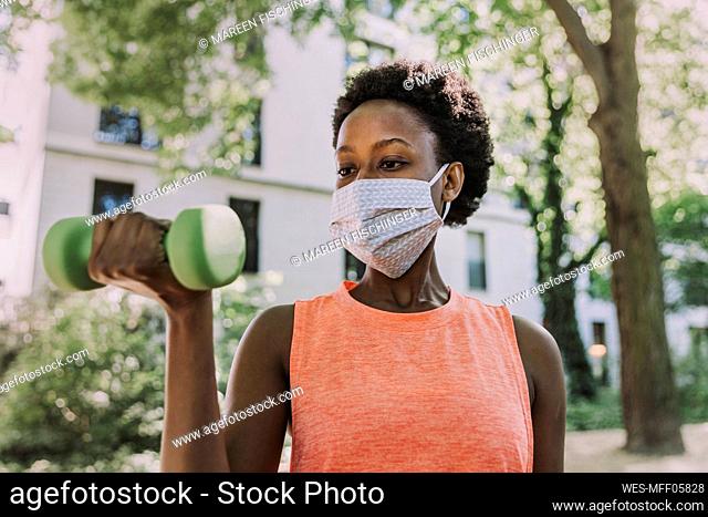 Portrait of woman wearing protective mask exercising with dumbbell outdoors