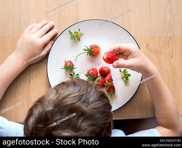 Little unrecognizable boy eats fresh strawberry with relish. Happy child eats organic strawberry at the kitchen table. Top view