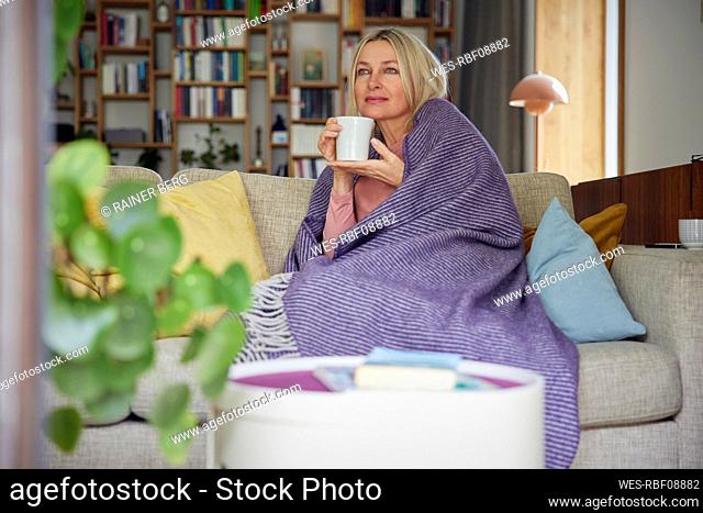 Blond woman wrapped in blanket holding coffee cup on sofa day dreaming at home