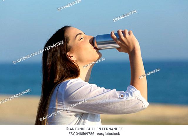 Side view portrait of a happy woman refreshing drinking soda sitting on a bench on the beach
