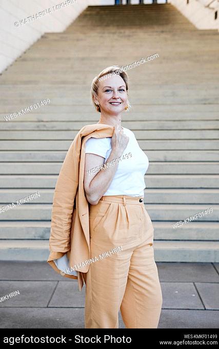 Smiling female entrepreneur holding blazer jacket while standing against staircase