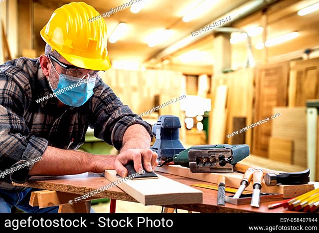 Carpenter worker at work in the carpentry workshop, wears helmet, goggles and surgical mask to prevent coronavirus infection