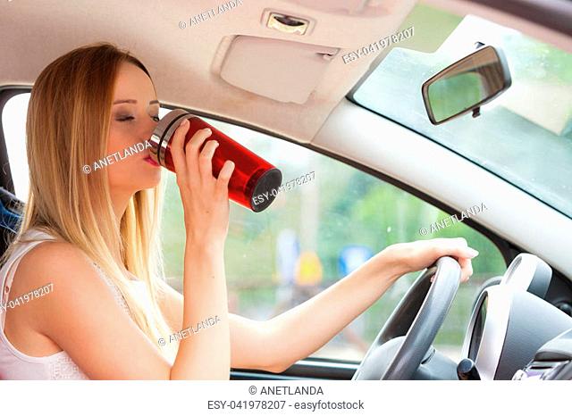 Take an energy boost. Young attractive woman sitting at the wheel drinking hot tea or coffee from vacuum flask thermos while driving the car