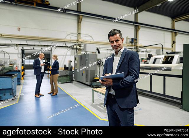 Portrait of a confident businessman using tablet in a factory with colleagues in background