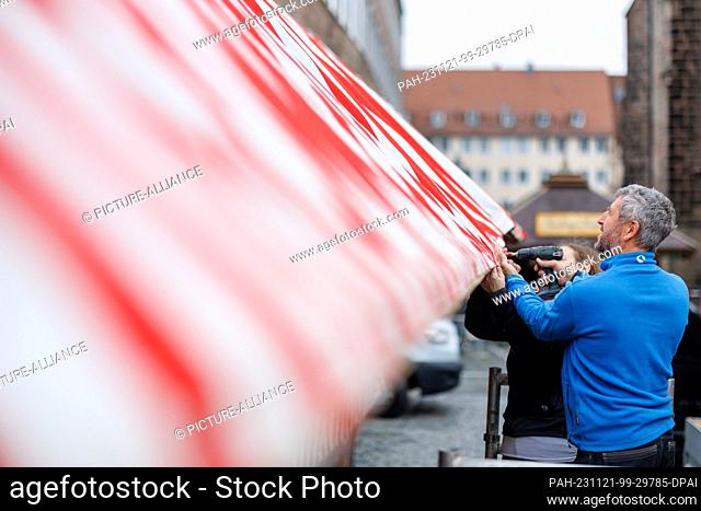 21 November 2023, Bavaria, Nuremberg: A man attaches a red and white striped roof tarpaulin to a stand during set-up work at the Nuremberg Christmas Market