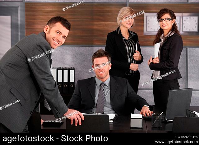 Business executive sitting at desk in office, looking at camera with team standing around
