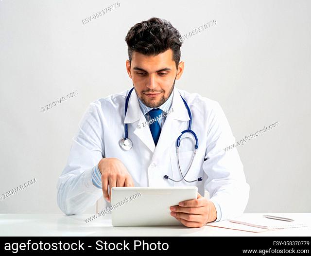 Young arab internist using tablet computer. Serious therapist in white medical gown with stethoscope sitting at desk. Modern digital technology in diagnosis and...