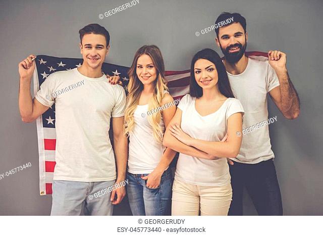 Beautiful young friends are holding an American flag, looking at camera and smiling, on gray background