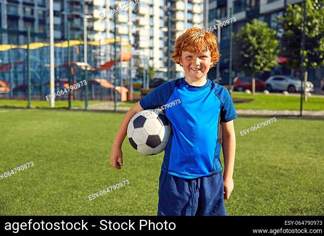 Cute redhead boy child wearing sportive uniform with ball portrait. Young football player at children soccer school concept