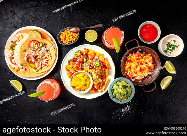 Mexican food, a flat lay overhead shot on a black background. Nachos, tacos, guacamole, shrimps, Paloma cocktails, limes and dips