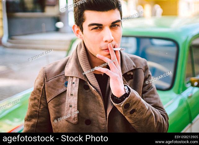 Portrait of a model young handsome sexy male brunette guy with dark skin Turkish Middle Eastern brunette posing smoking a cigarette near old retro car on the...