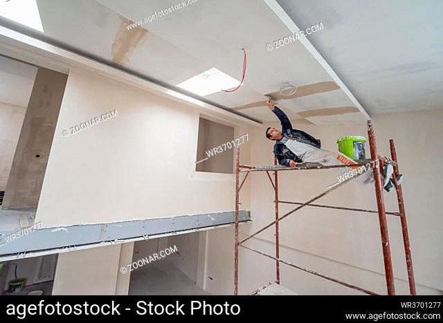 young professional construction worker using scaffold while plastering on gypsum ceiling inside the new big modern two levels apartment