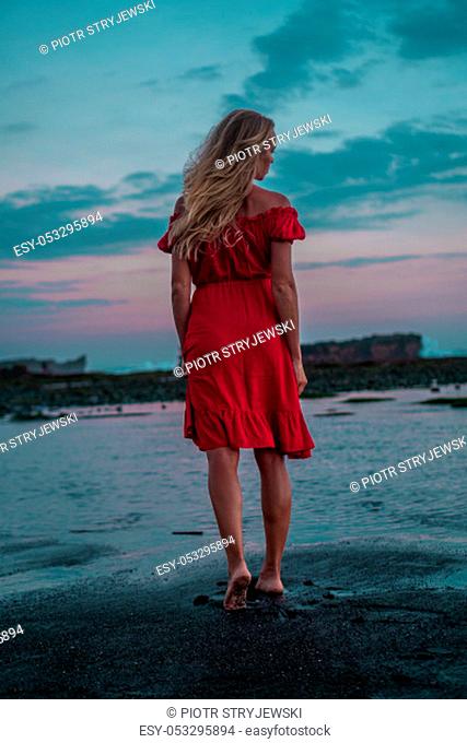 Back view of beautiful sensual woman in red dress posing on the black sand beach during sunset