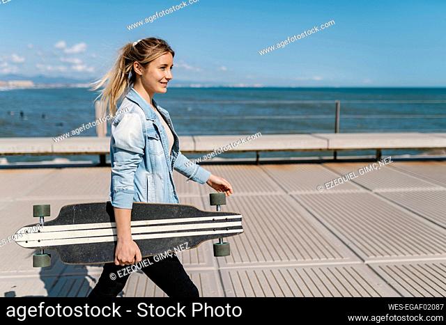 Smiling woman with longboard walking on pier during sunny day