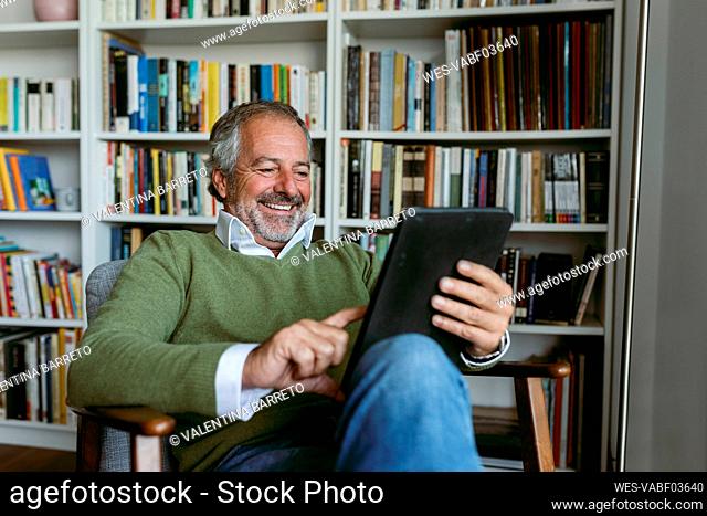 Smiling man using digital tablet while sitting against bookshelf at home