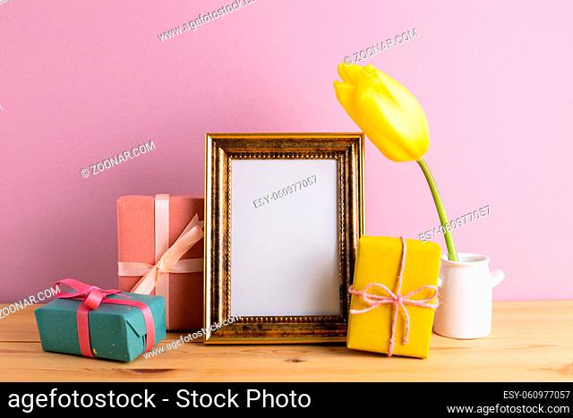 Photo frame with gift boxes and yellow tulip on wooden table with pink background. Anniversary concept, floral arrangement, copy space
