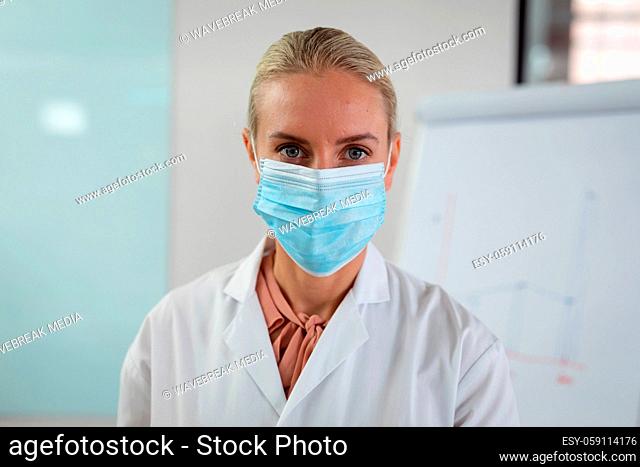 Portrait of caucasian female doctor wearing face mask sitting in hospital office looking to camera