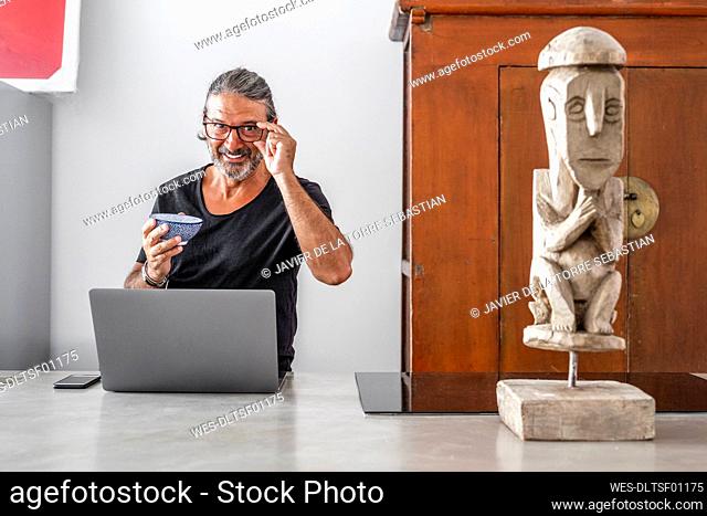 Smiling man with bowl using laptop while standing in kitchen